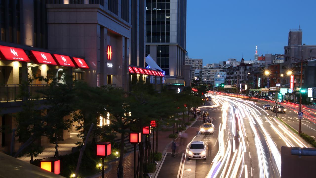 Caesar Metro Taipei Hotel Bagian luar foto The photo shows a city scene during twilight. In the foreground, there is a busy road with streaks of light from moving vehicles, suggesting it's a lively area. On the sides of the street, red lanterns or light fixtures are visible, adding to the amb