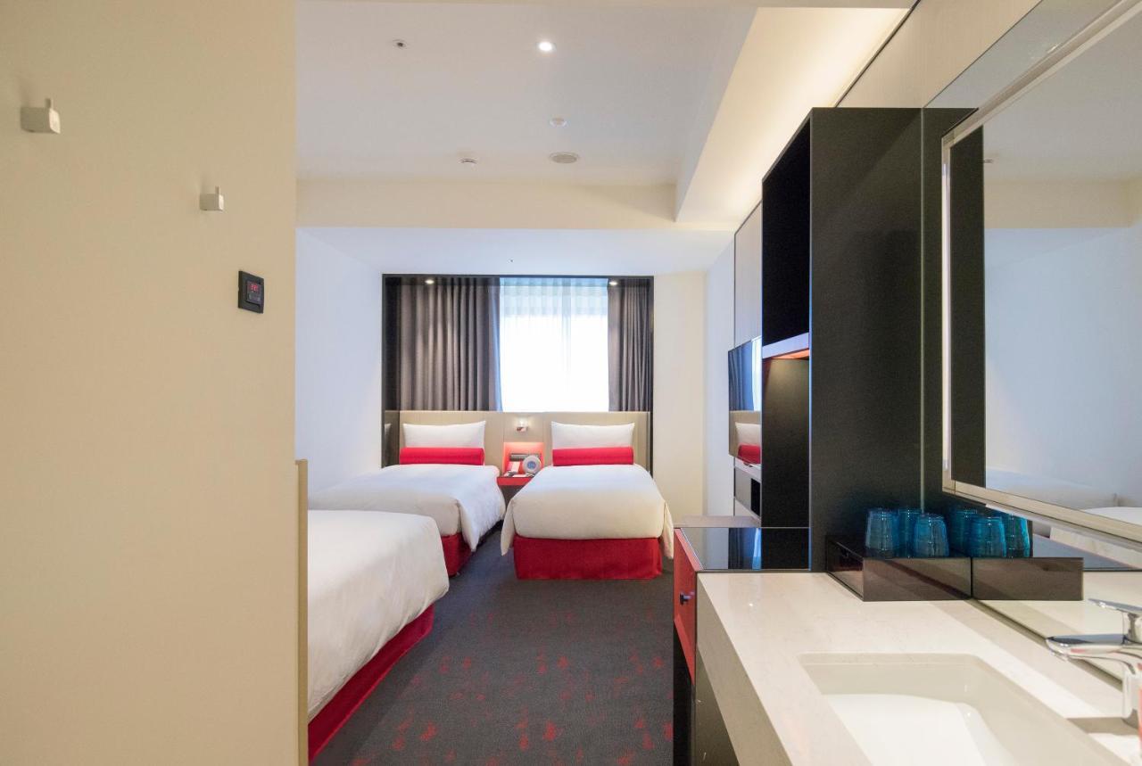 Caesar Metro Taipei Hotel Bagian luar foto The photo shows the interior of a hotel room. It features two single beds with white linens and red accents. There is a window with sheer curtains allowing natural light to enter the room. The floor is carpeted, and the decor is modern and minimalist